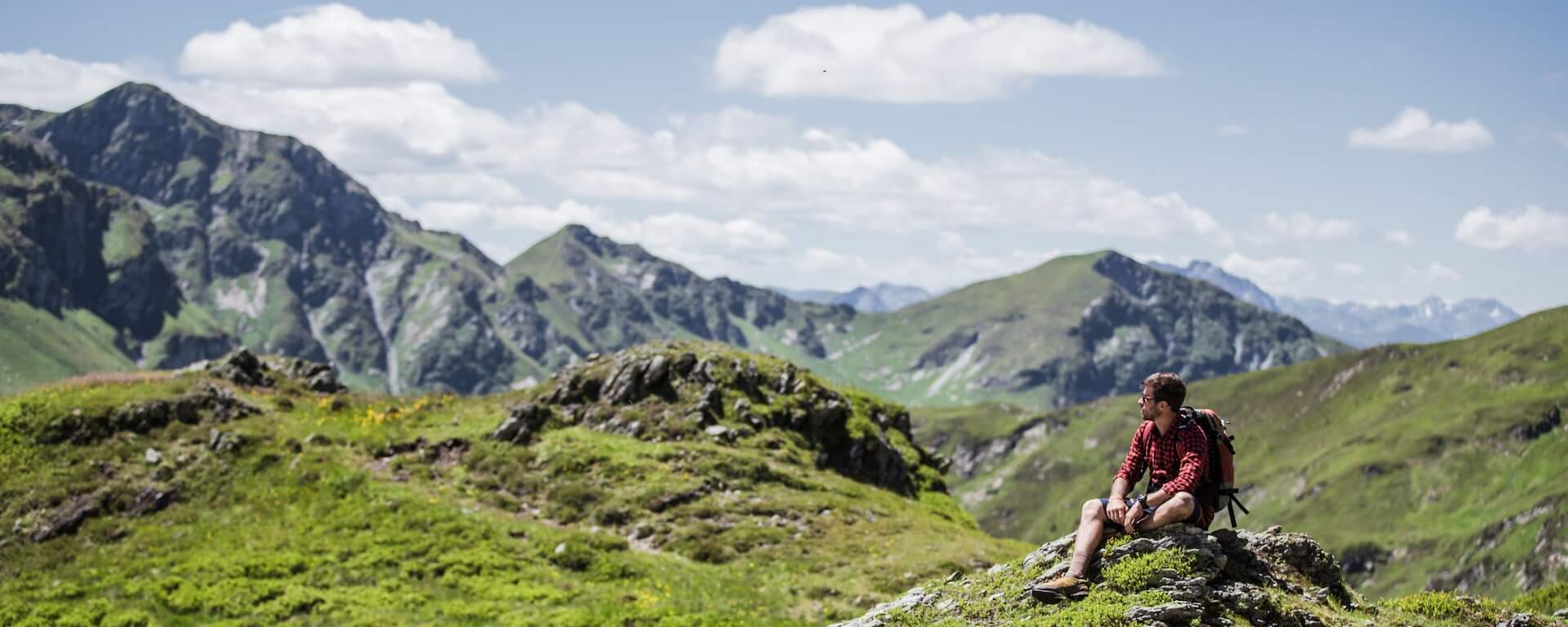 Wanderer ruht auf einem Felsen in den Alpen, umgeben von grünen Hügeln und Bergen.