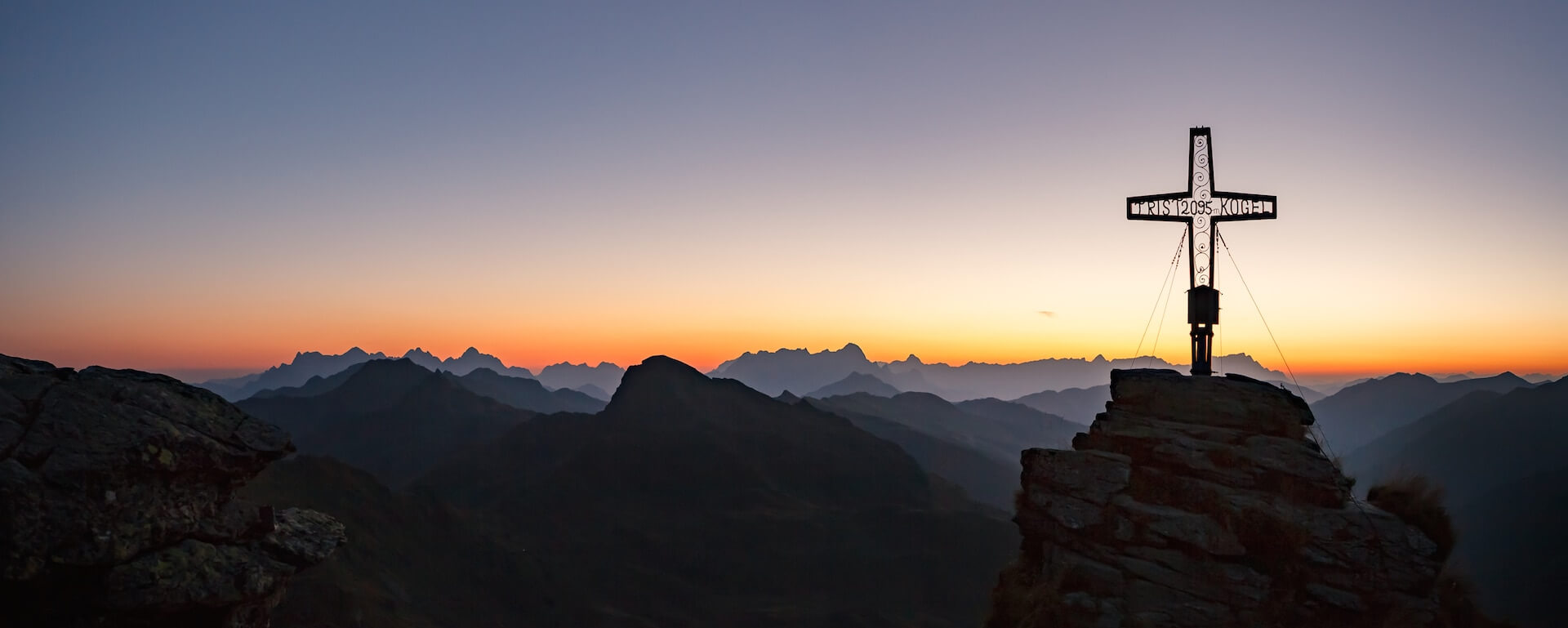 Sonnenuntergang über den Alpen mit Bergkreuz und beeindruckender Landschaft.